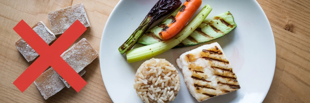 Assiette avec légumes grillés, poisson, riz et indication de modération des sucreries avec une croix rouge, symbolisant une alimentation saine et équilibrée. Fond en bois chaleureux.